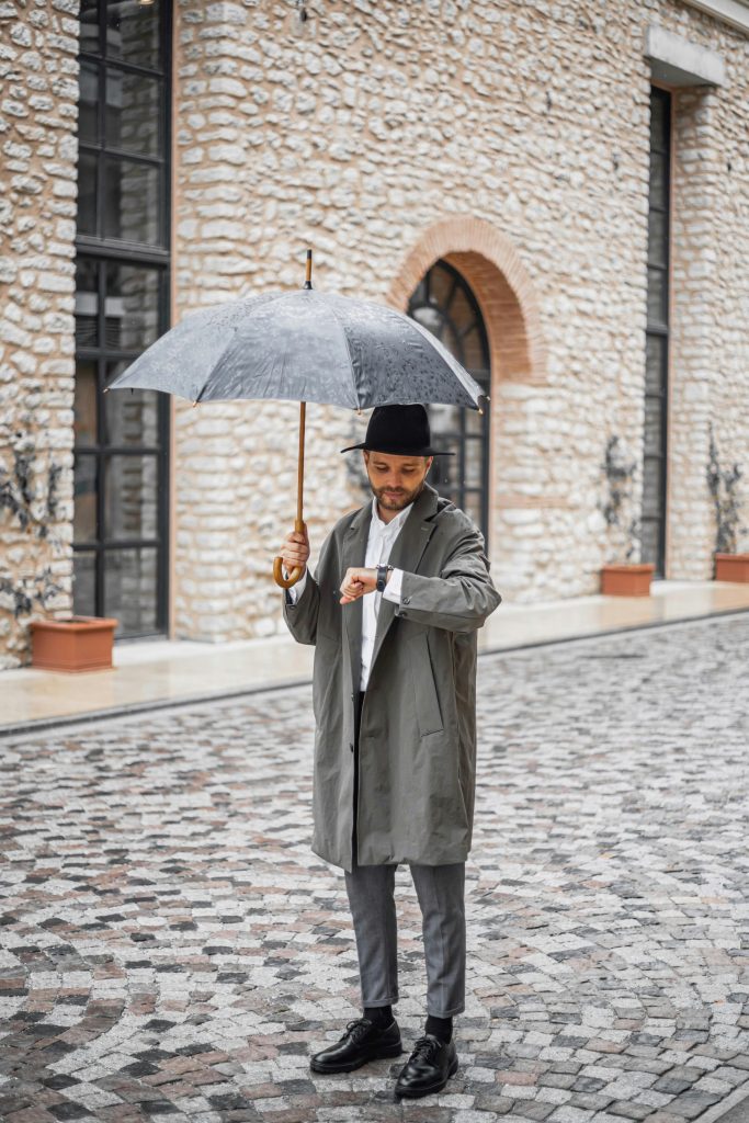 A man in a stylish outfit checks his watch while holding an umbrella on a cobblestone street in Istanbul.