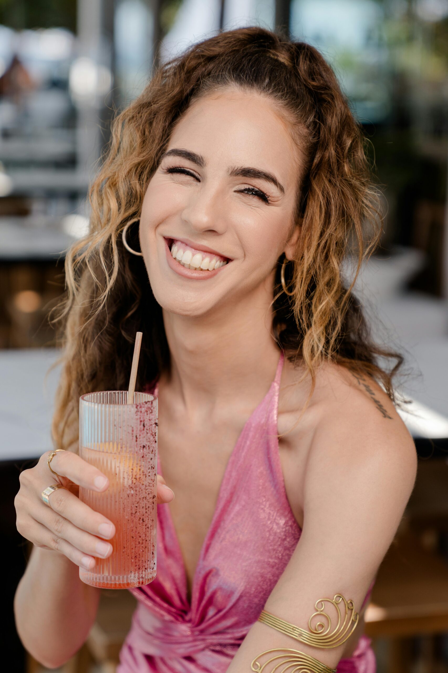 Young woman in a pink dress smiling while holding a refreshing drink in a stylish glass outdoors.