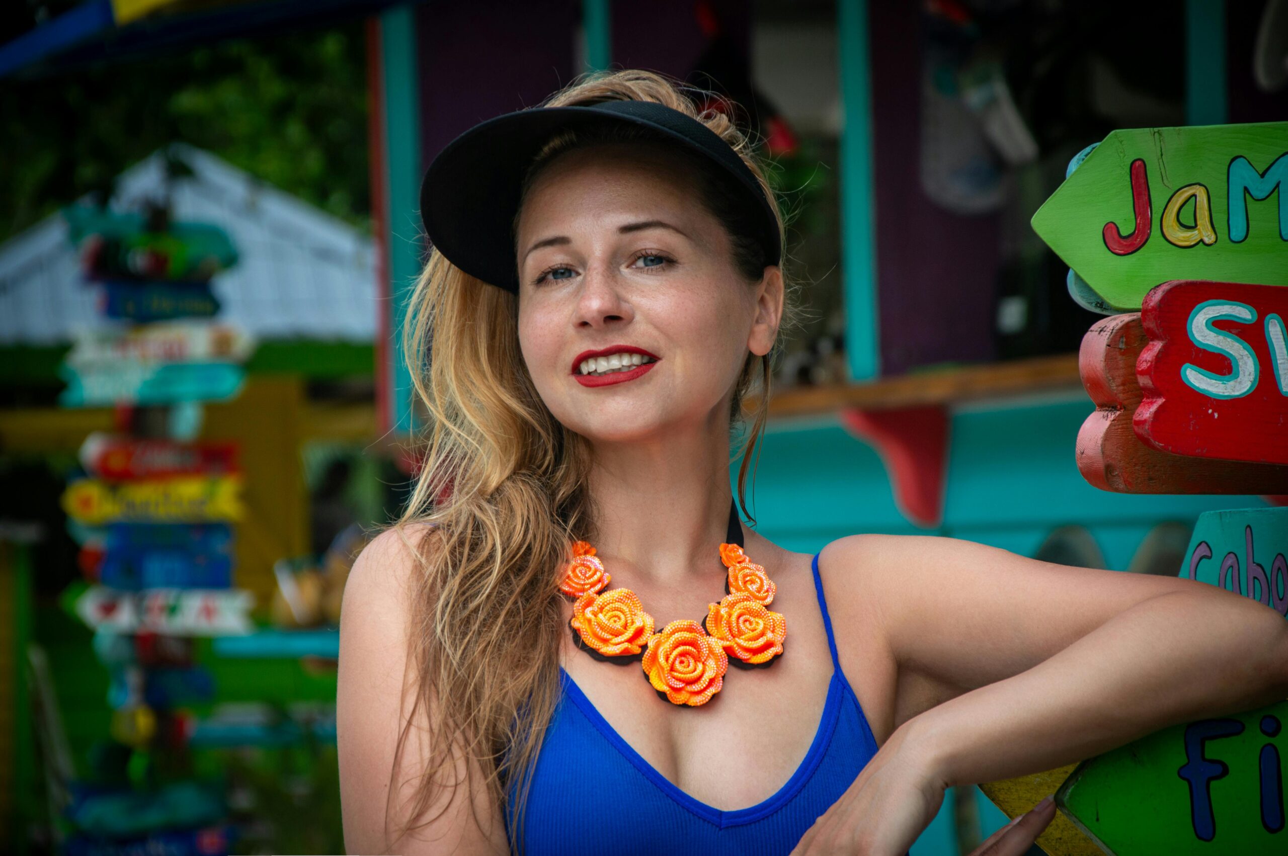 Cheerful woman in vibrant jewelry and visor smiles in colorful tropical setting.